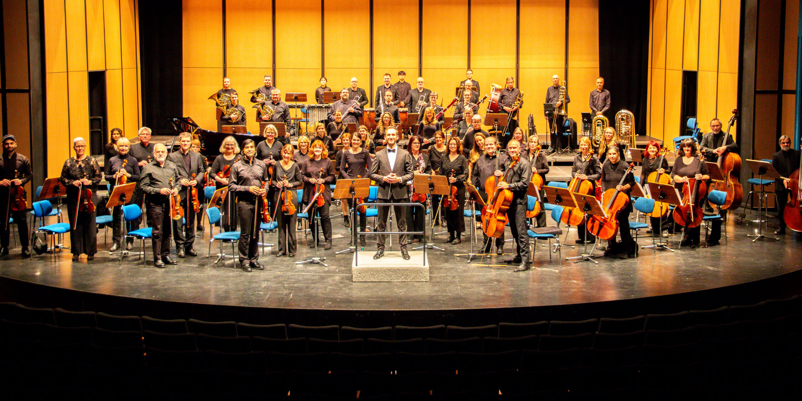 Orchester mit vielen Musikern und Instrumenten auf einer Bühne mit gelbem Hintergrund