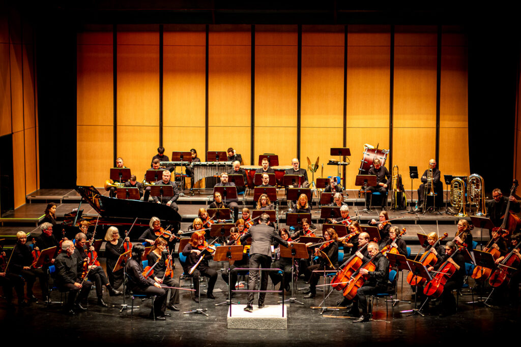 Orchester spielt auf Bühne mit Holzwand, Dirigent leitet von vorne