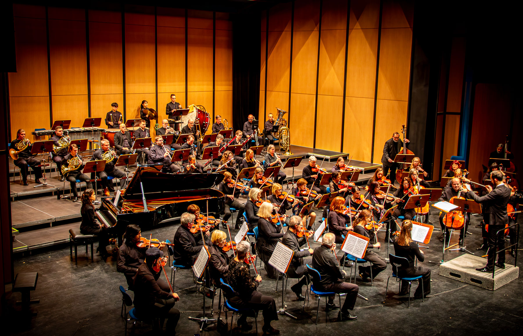 Orchester mit Dirigent und Flügel auf Bühne, von vorne, warme Beleuchtung